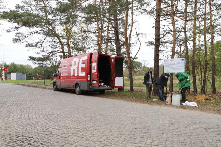 Trzech pracowników firmy montażowej  wkopują tablicę na poboczu drogi. W tle duży czerwony samochód transportowy