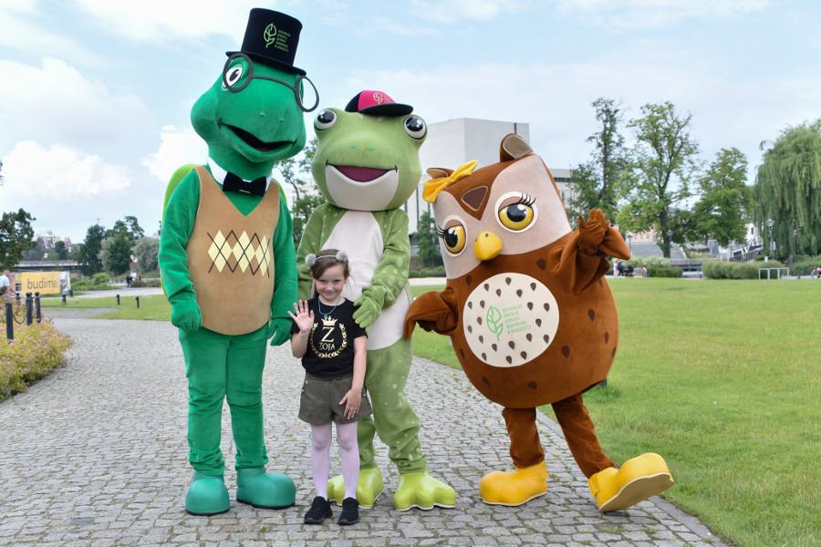 Zywa maskotki: żółw, żaba i sowa, pozują do zdjęcia z machającą małą dziewczynką