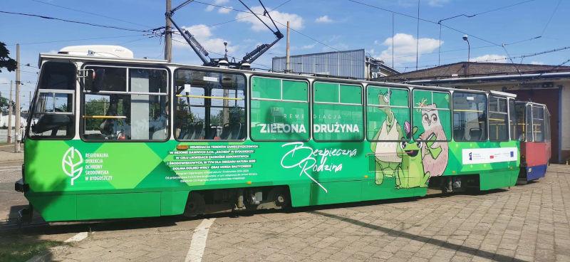 W zajezdni stoi tramwaj. Pierwszy wagon jest zielony i widać na nim logotypy i opis projektu